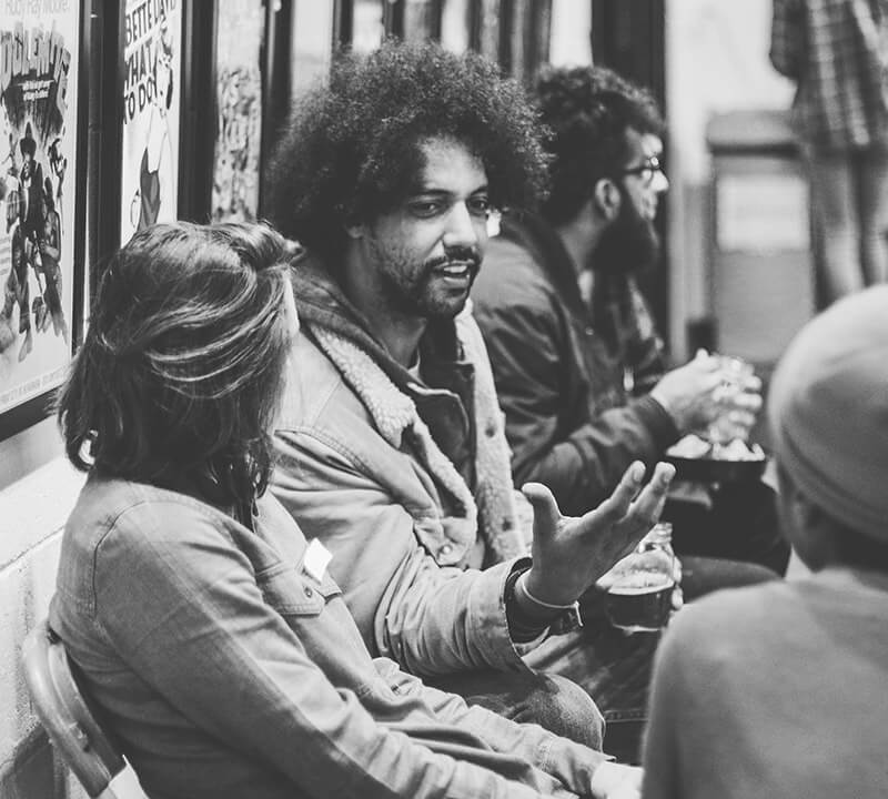 A black man smiling as he speaks expressively with others sitting near him at a Two Bettys employee party.