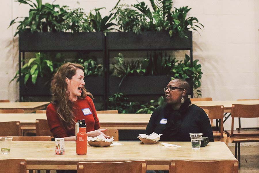 Two women, one white, one black, talking animatedly at a table in front of a wall of green plants.