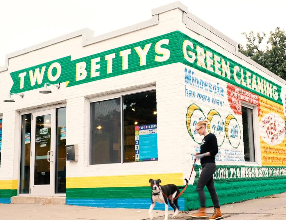 A person in black clothes and tan boots walking a brown and white dog outside of Two Bettys Refill Station.