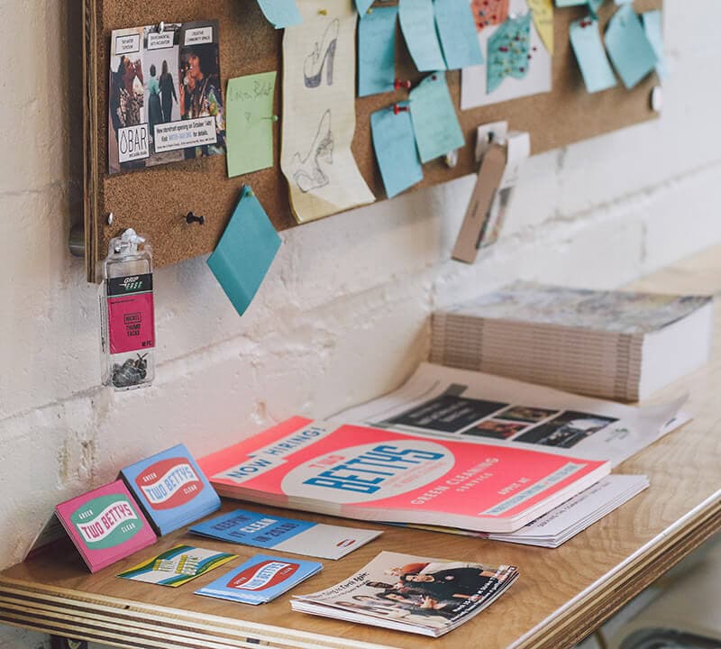 Two Bettys Green Cleaning business cards and posters on a wooden surface.