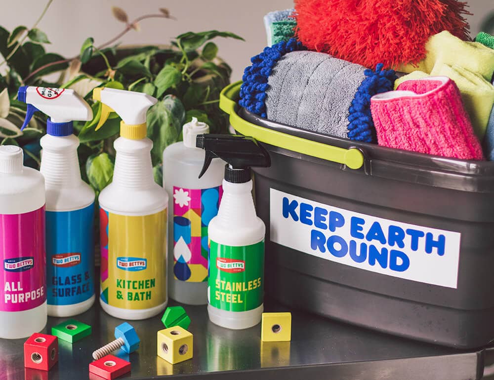 Two Bettys Green Cleaning bottles of cleaning supplies, a bucket full of cleaning cloths with a "Keep Earth Round" sticker on the front