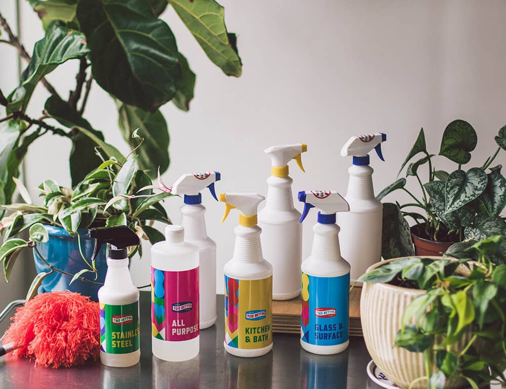 Seven bottles of Two Bettys Green Cleaning products on a metal surface surrounded by green plants.