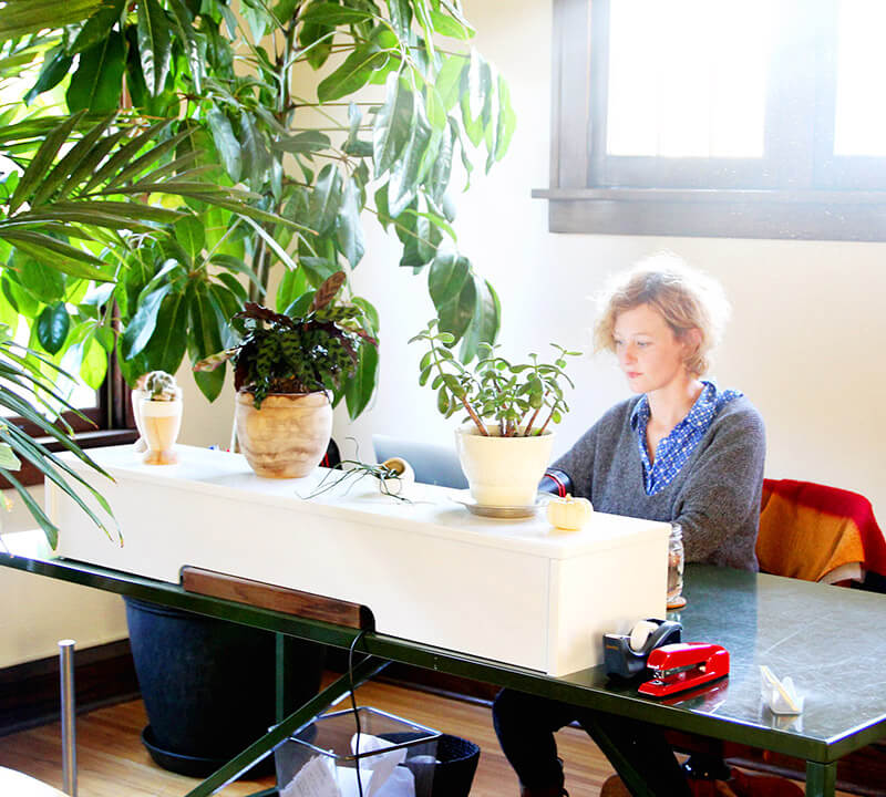 A white woman sitting behind a large desk among green plants at Two Bettys Green Cleaning HQ.