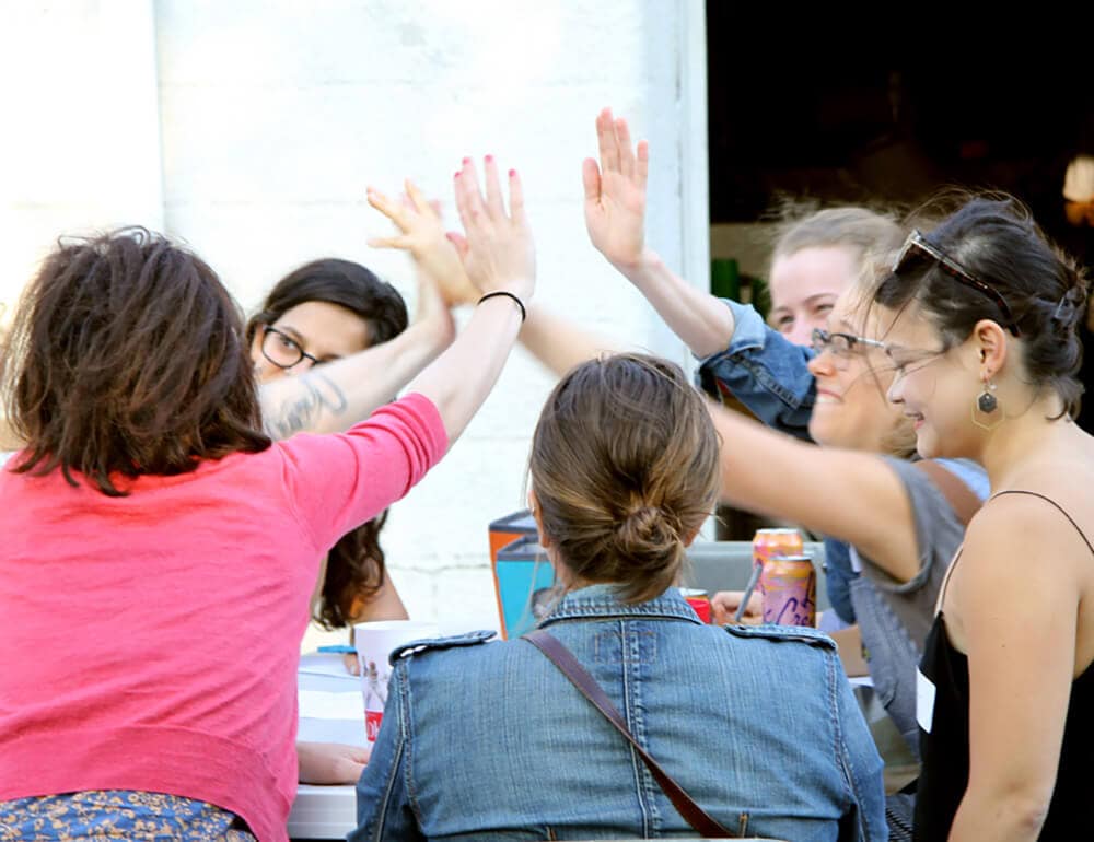 A group of Two Bettys Green Cleaning cleaners sitting around a table and high fiving.