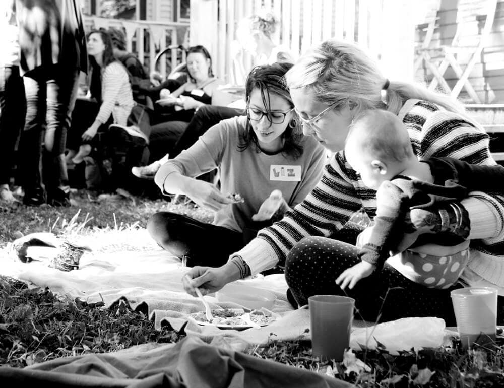 Two women, one wiht a baby on her hip, sitting and eating picnic style