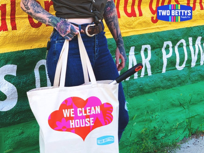 Two Bettys tote bag with "WE CLEAN HOUSE" printed in a bright red and pink shape, being held by a heavily tattooed person with long pink nails