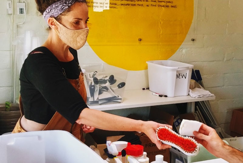 A person wearing a black shirt and overalls and a tan face mask handing off a red and white scrub brush to another person outside of the frame.