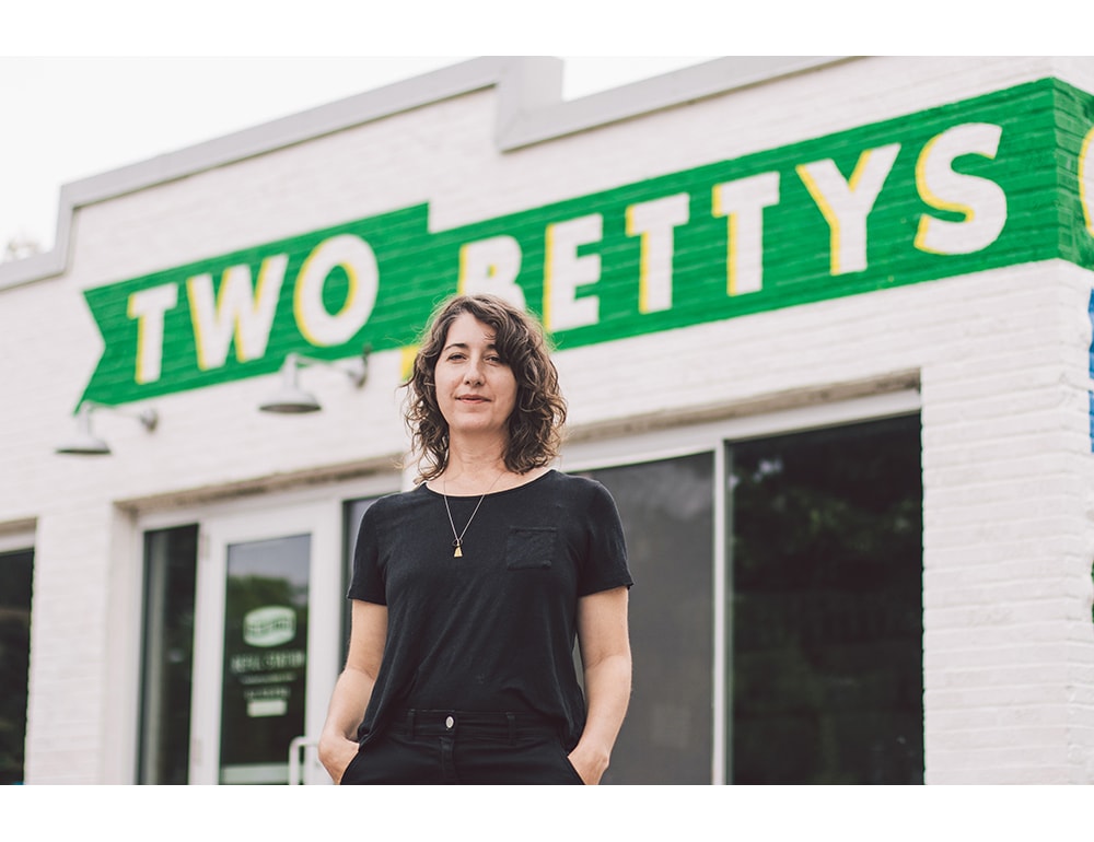 A white woman with wavy brown hair standing in front of the Two Bettys building