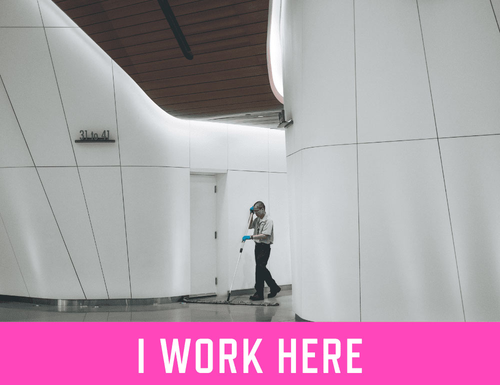 A janitor cleaning the floors in a bright white modern office building setting with text "I WORK HERE" at the bottom. Photo by Verne Ho.