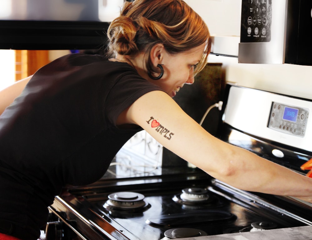 A white woman with her hair in buns and "I (heart) MPLS" tattooed on the back of her arm cleaning a black enamel gas stove top.